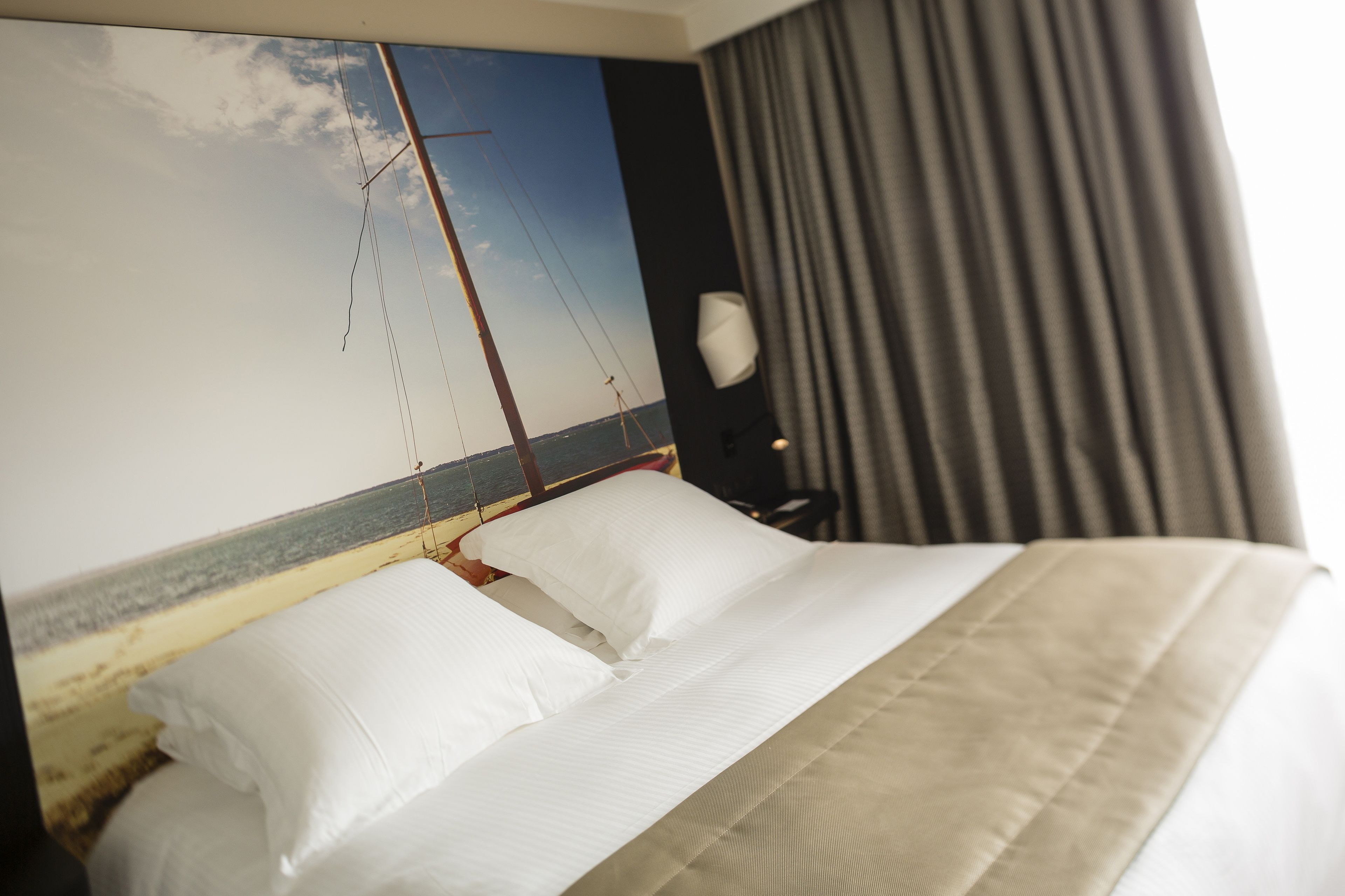 A hotel room with a bed, a headboard with a painting of a sailboat, and a large window with curtains.