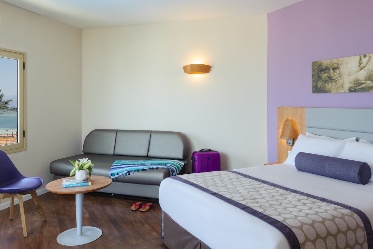A hotel room with a bed, a sofa, a small table with a plant, and a window with a view of the beach.
