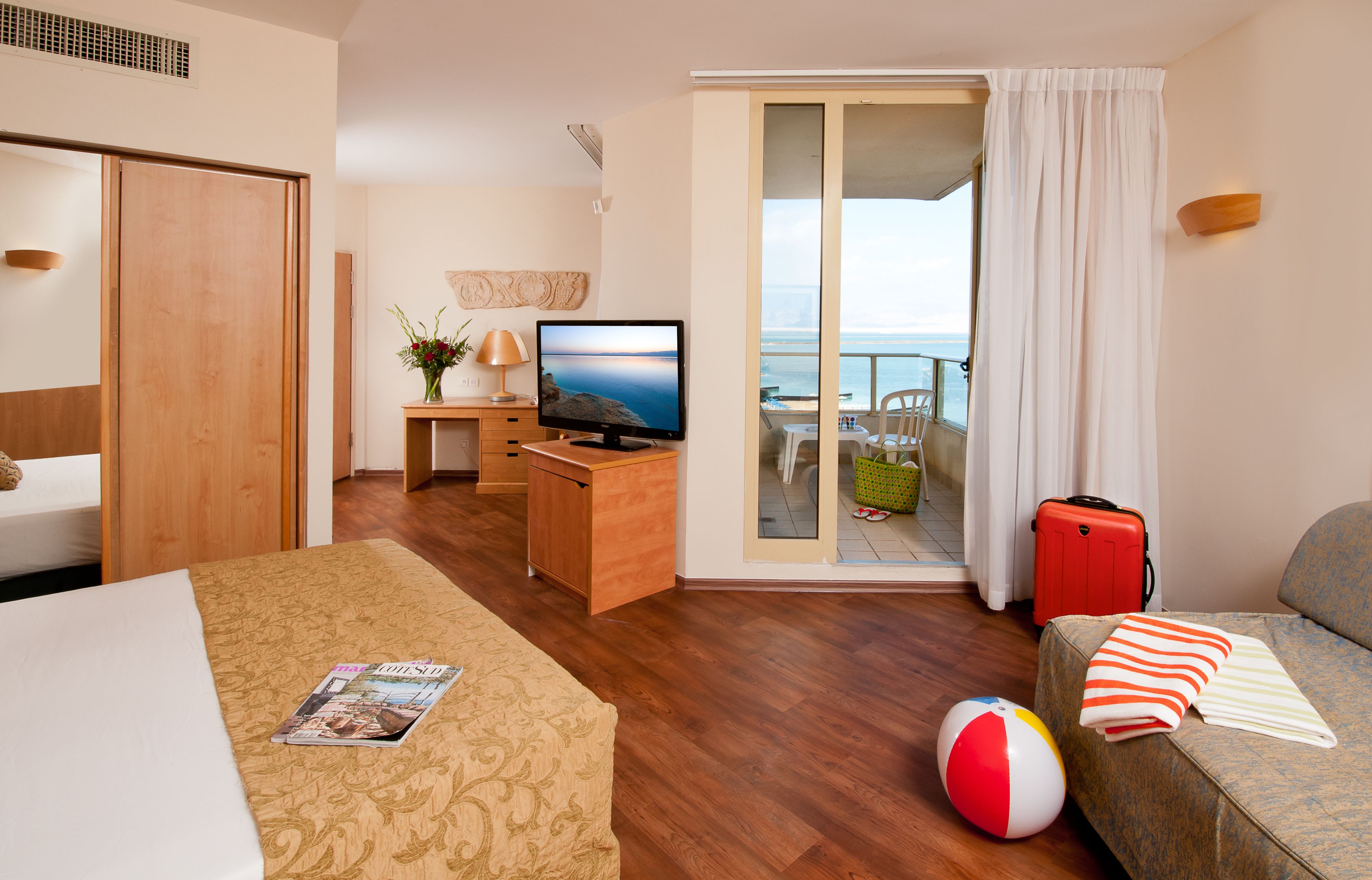 Ett hotellrum med en säng, ett skrivbord med en dator, en tv, en balkong med utsikt över havet och en röd resväska.