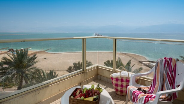 A balcony with a view of the beach and ocean, a wicker basket with fruit, a checkered beach chair, and a white chair with a striped towel. - Leonardo Plaza Dead Sea (Tamar)