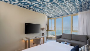 A hotel room with a bed, a sofa, a desk with a TV, and a large window with a view of the sea. - Leonardo Plaza Dead Sea (Tamar)