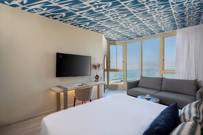 A hotel room with a bed, a sofa, a desk with a TV, and a large window with a view of the sea. - Leonardo Plaza Dead Sea (Tamar)