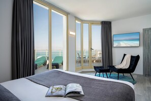 A hotel room with a large bed, a view of the ocean, a balcony with outdoor seating, and a painting on the wall.