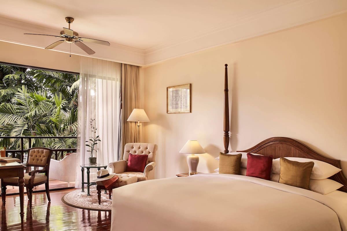 A hotel room with a large bed, a sitting area with a chair and table, a balcony with plants, and a ceiling fan.