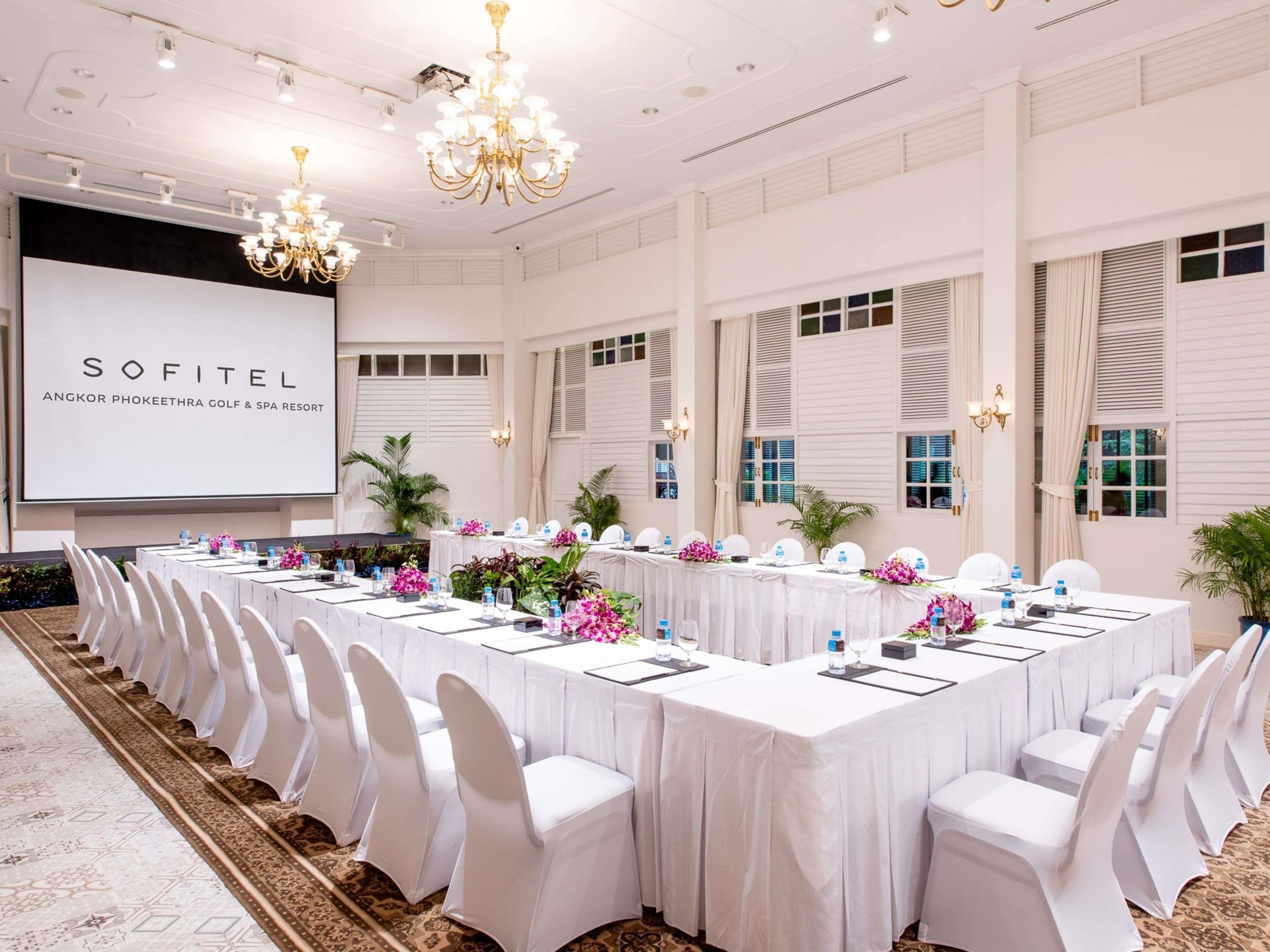 Kokoushuone Sofitel Angkor Phokeethra Golf & Spa -lomakeskuksessa, jossa on pitkä pöytä, valkoiset tuolit ja projektorikangas, johon on heijastettu lomakeskuksen nimi.