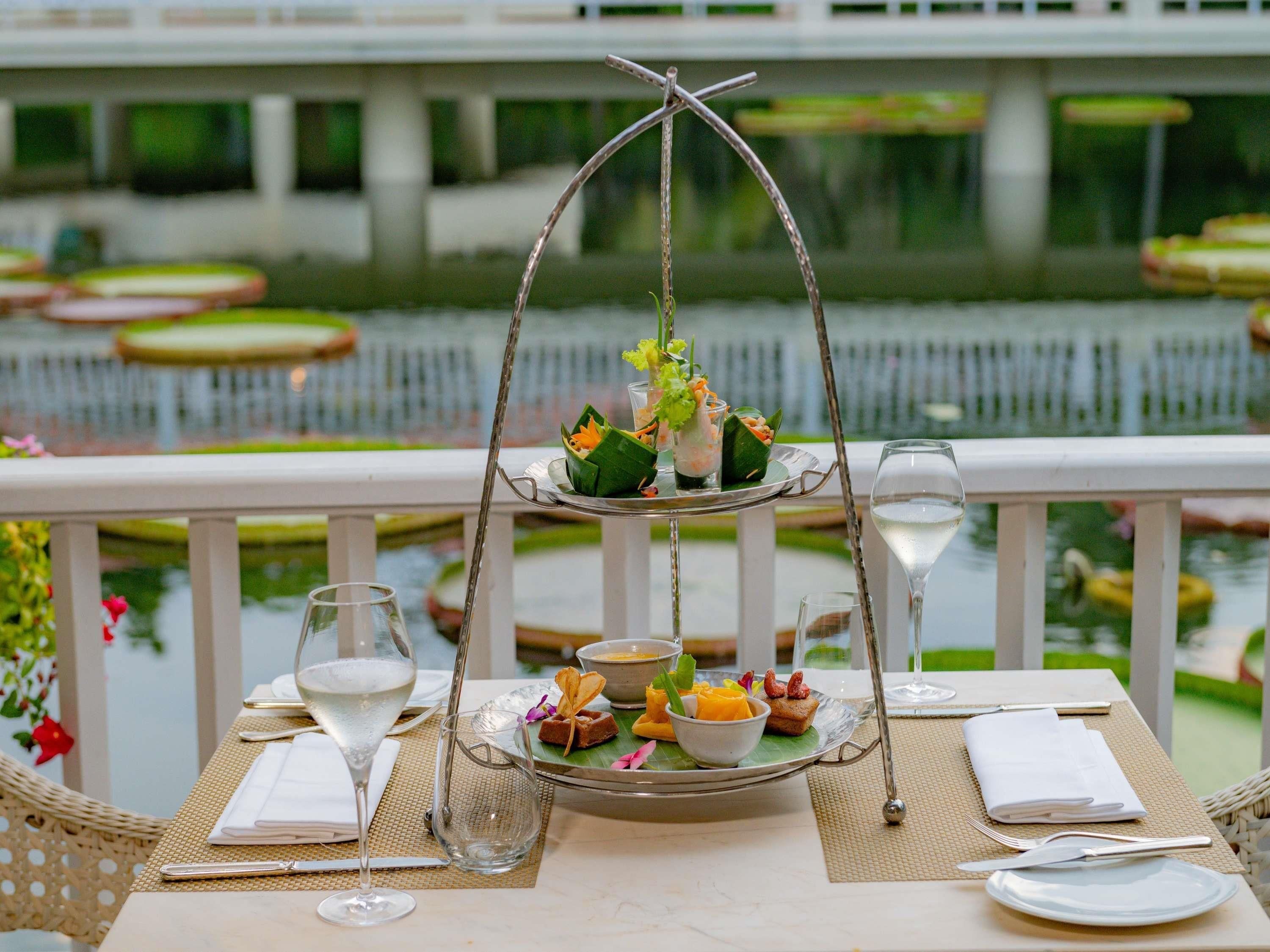 A dining table with a three-tiered serving tray, a glass of white wine, and a view of a water body with lily pads.