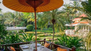 A dining table set with plates, glasses, and cutlery, surrounded by wicker chairs and orange cushions, with a view of a garden and water feature.