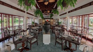 A spacious dining area with multiple tables and chairs, large windows, and hanging plants. - Sofitel Angkor Phokeethra Golf & Spa Resort (Siem Reap)
