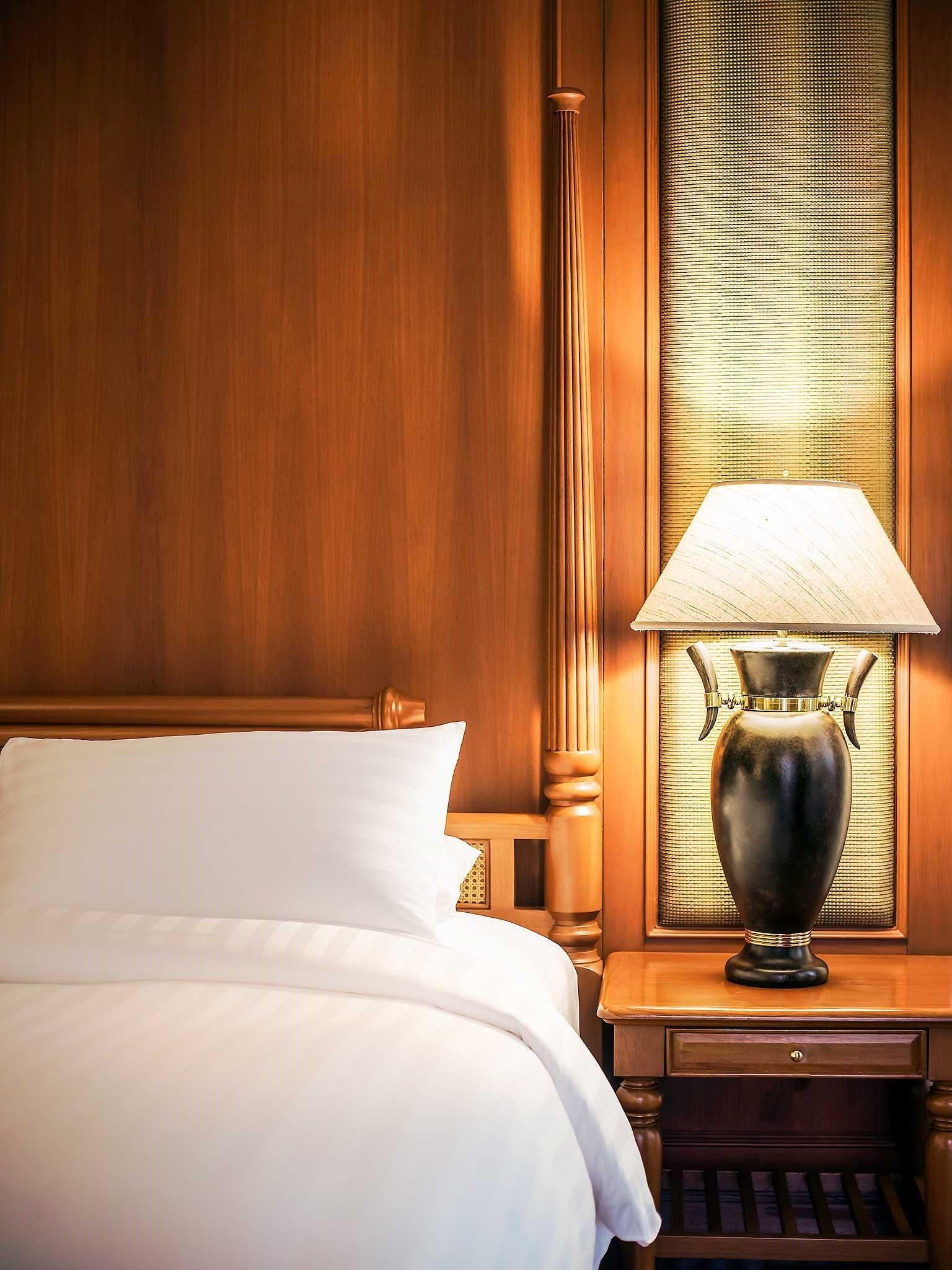 A hotel room with a wooden headboard, a bed with white bedding, a nightstand with a lamp, and a window with blinds.