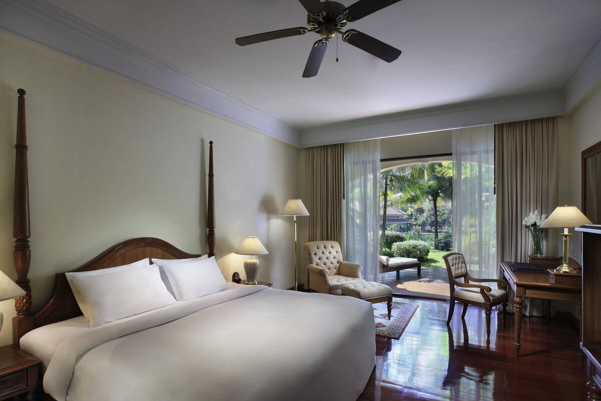 A hotel room with a large bed, a desk, a chair, a ceiling fan, and a view of the outdoors.