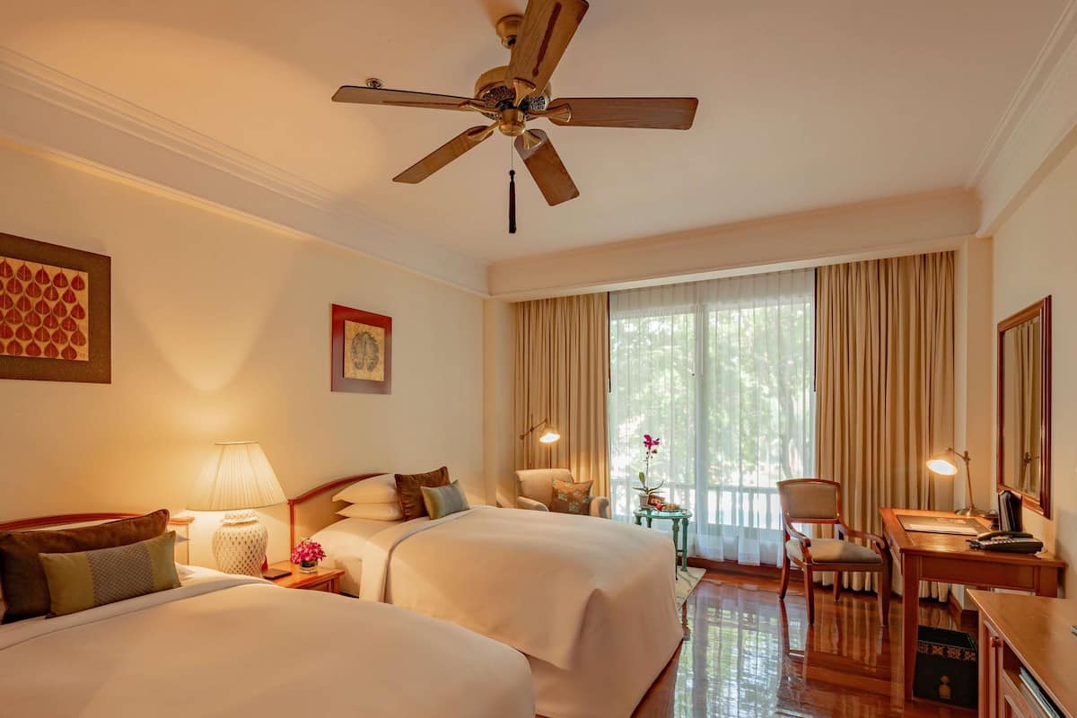 A hotel room with two beds, a desk, a chair, a ceiling fan, and a large window with curtains.