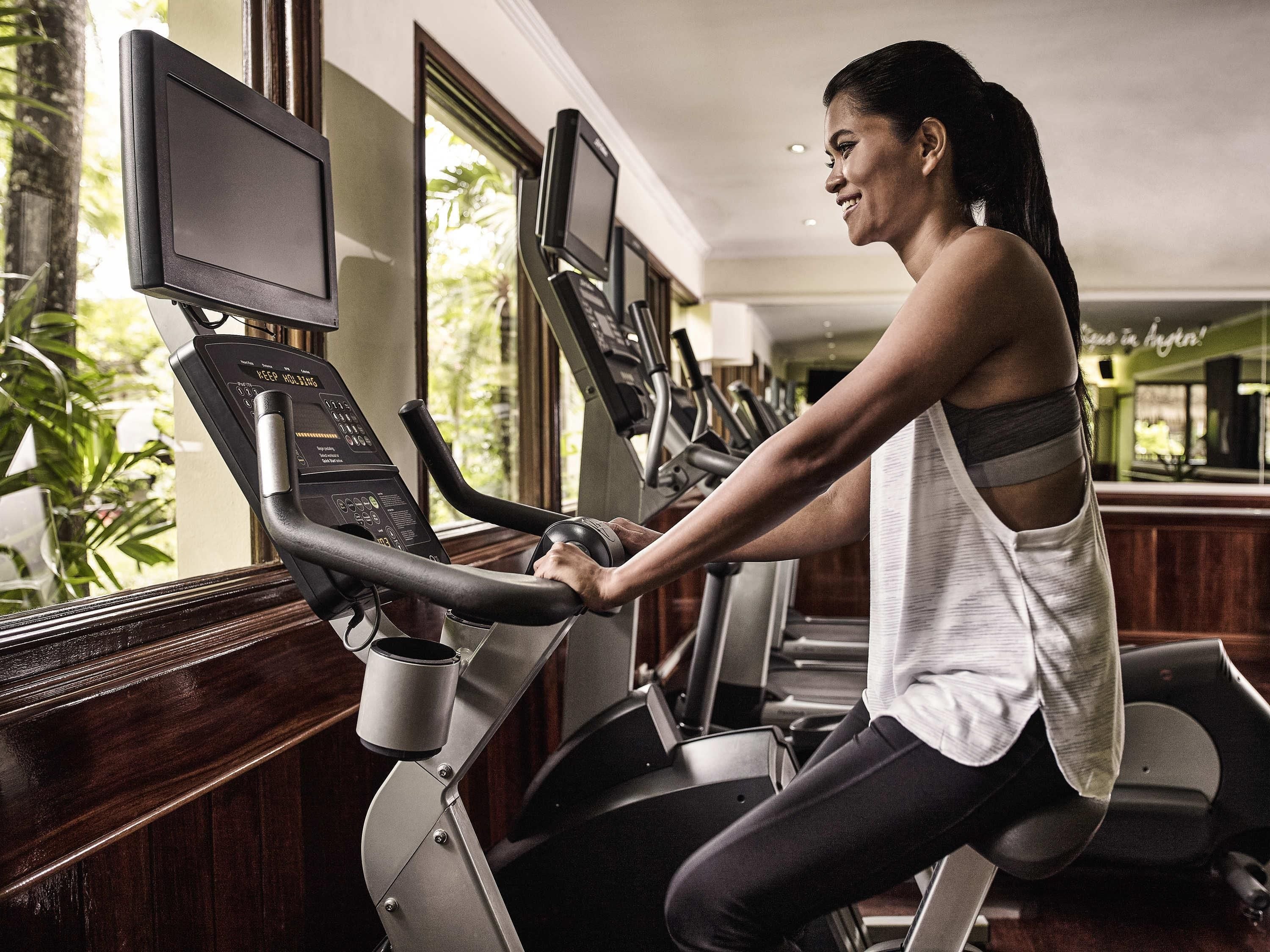 A woman using an elliptical machine in a gym with multiple treadmills and a large window providing a view of greenery outside.
