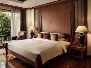 A large bed with a wooden headboard, two bedside tables with lamps, a chair, and a view of greenery through the window.