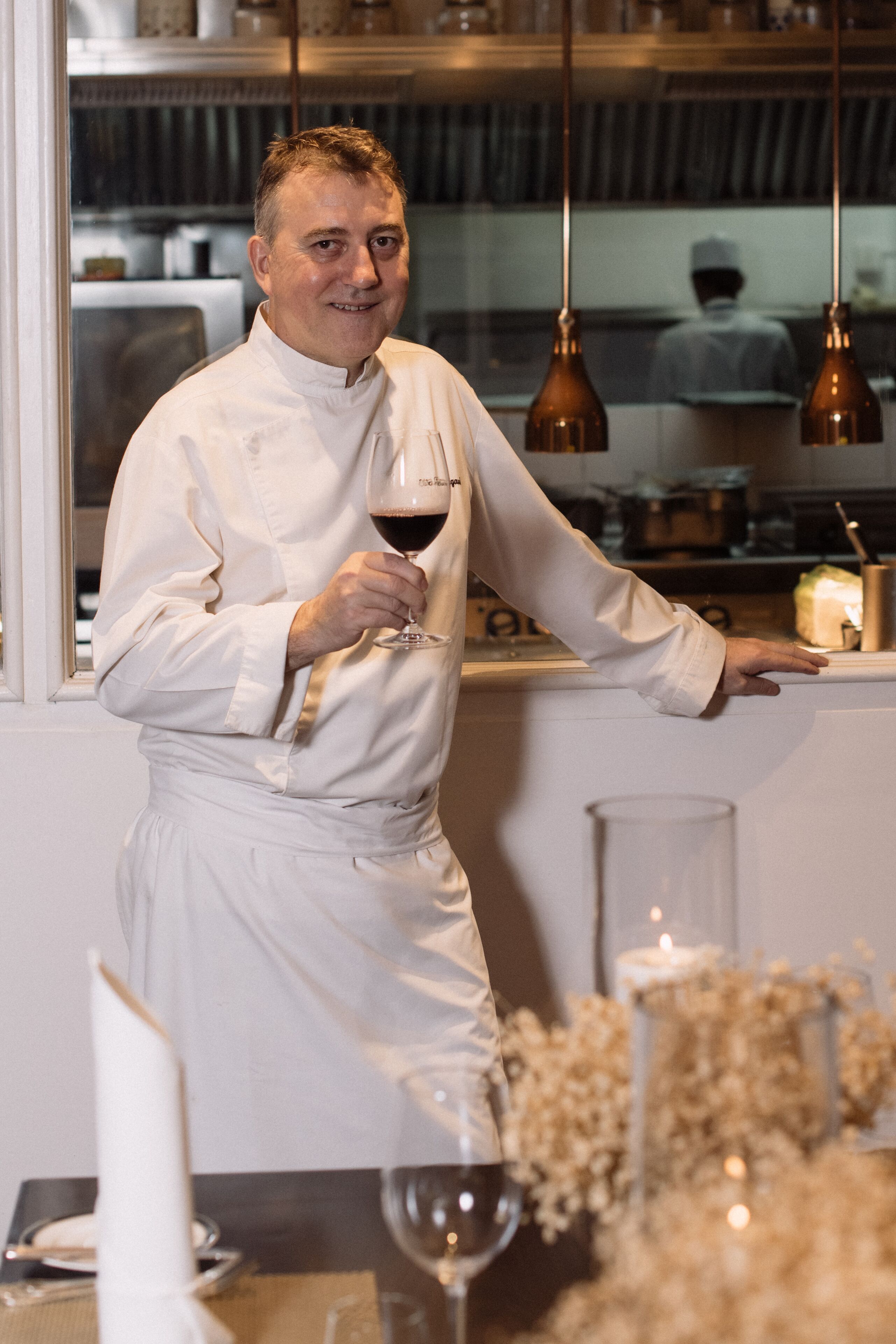 A chef in a white uniform holding a wine glass, standing in a kitchen with hanging lights and a window.