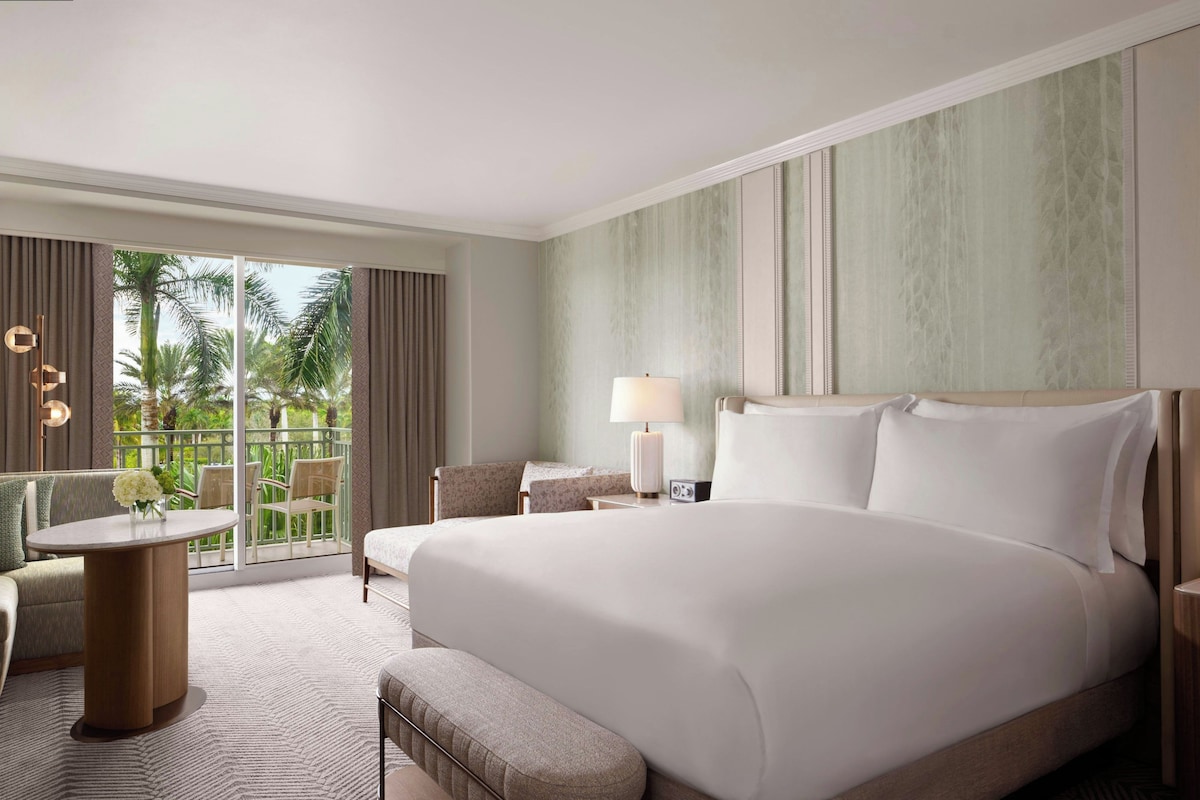 Quarto de hotel com uma cama grande, área de estar com mesa e cadeiras, luminária e vista para palmeiras pela janela.