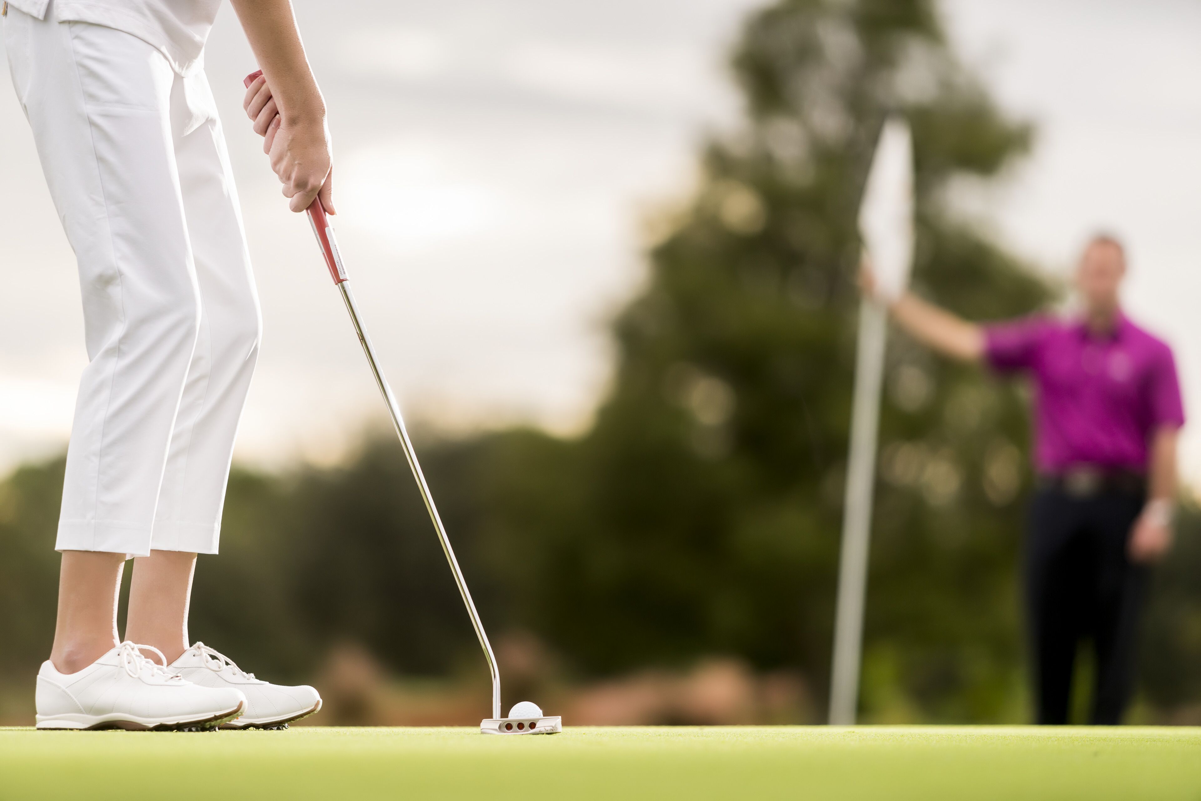 A person in white attire preparing to put a golf ball on a green, with a golf club in hand.