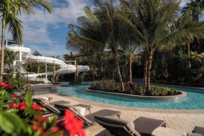 A pool area with lounge chairs, a water slide, palm trees, and a clear sky.