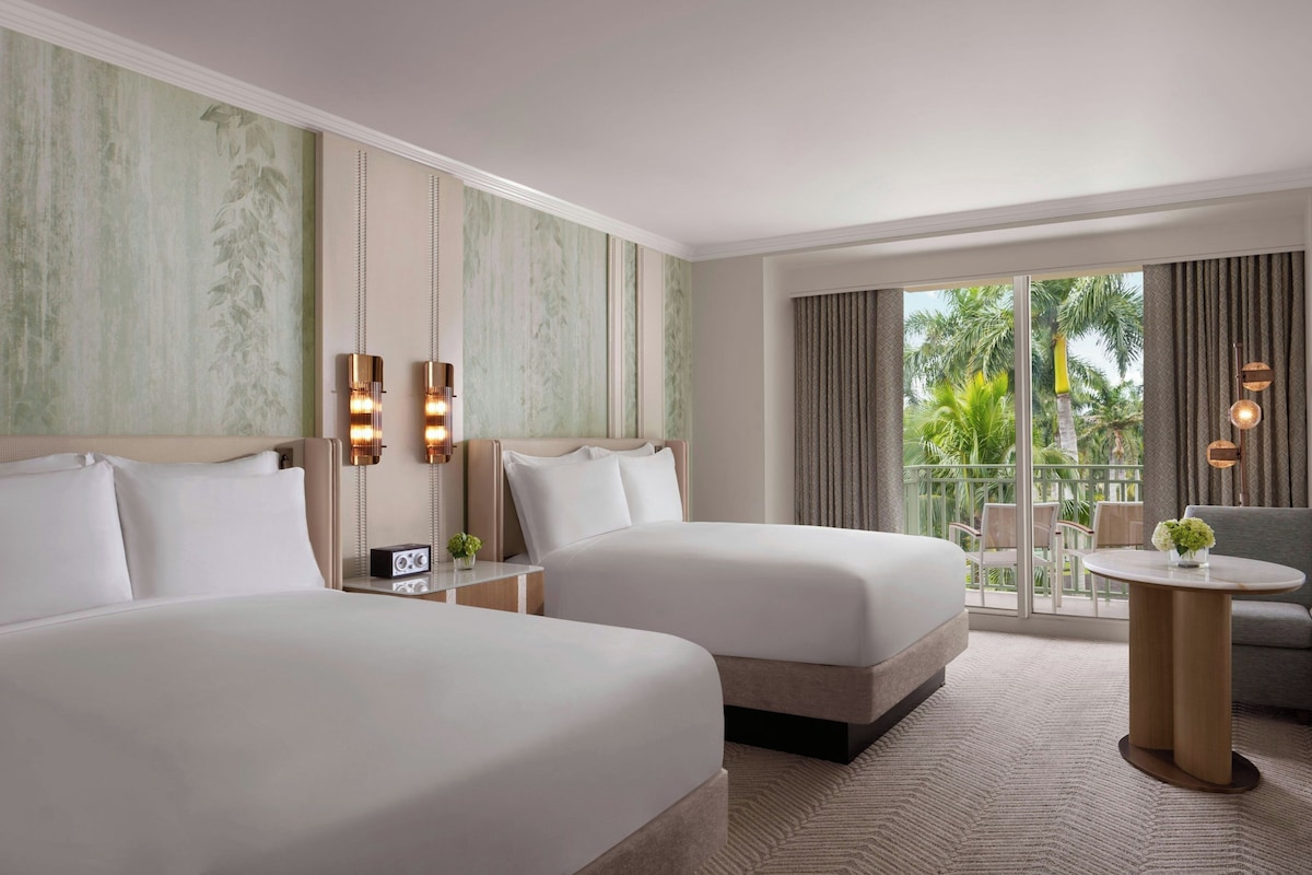 Quarto de hotel com duas camas, área de estar com mesa e cadeiras, varanda com vista para palmeiras e uma janela grande com cortinas.