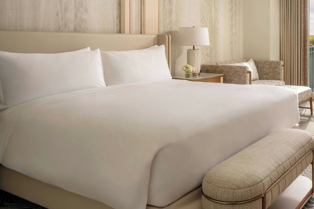 A large bed with white linens, a bedside table with a lamp, and a chair.
