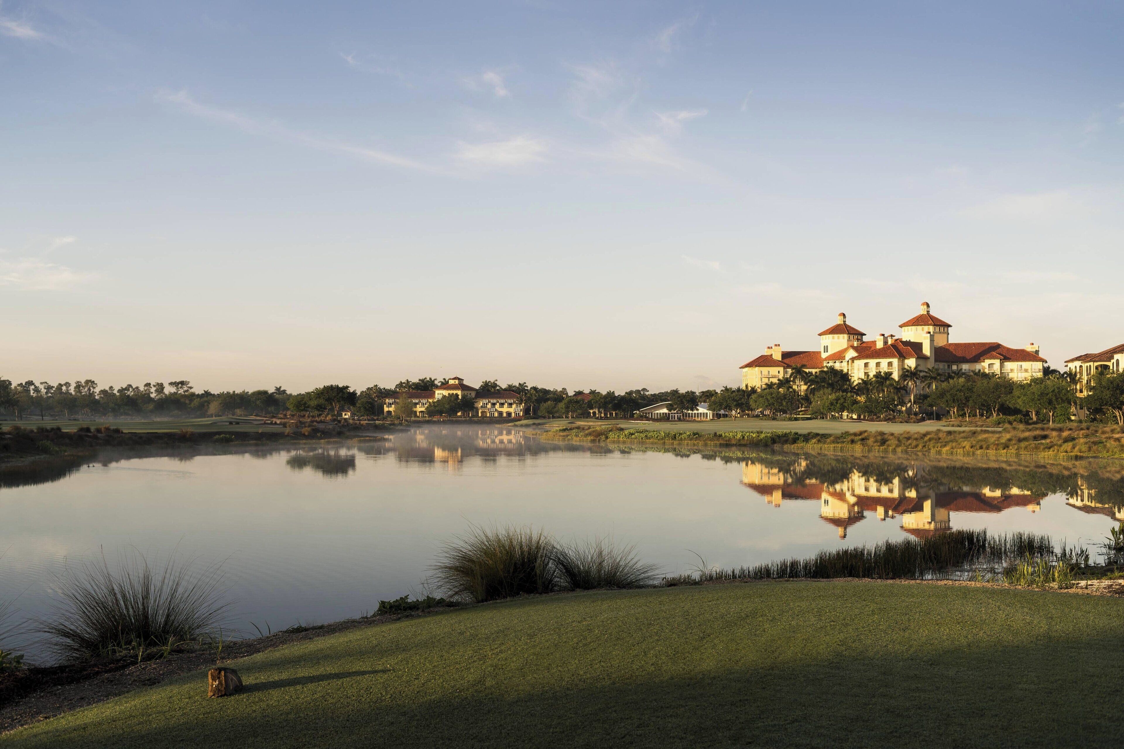 Una vista tranquila de un lago con un campo de golf, una extensa propiedad con varias construcciones y un cielo despejado.