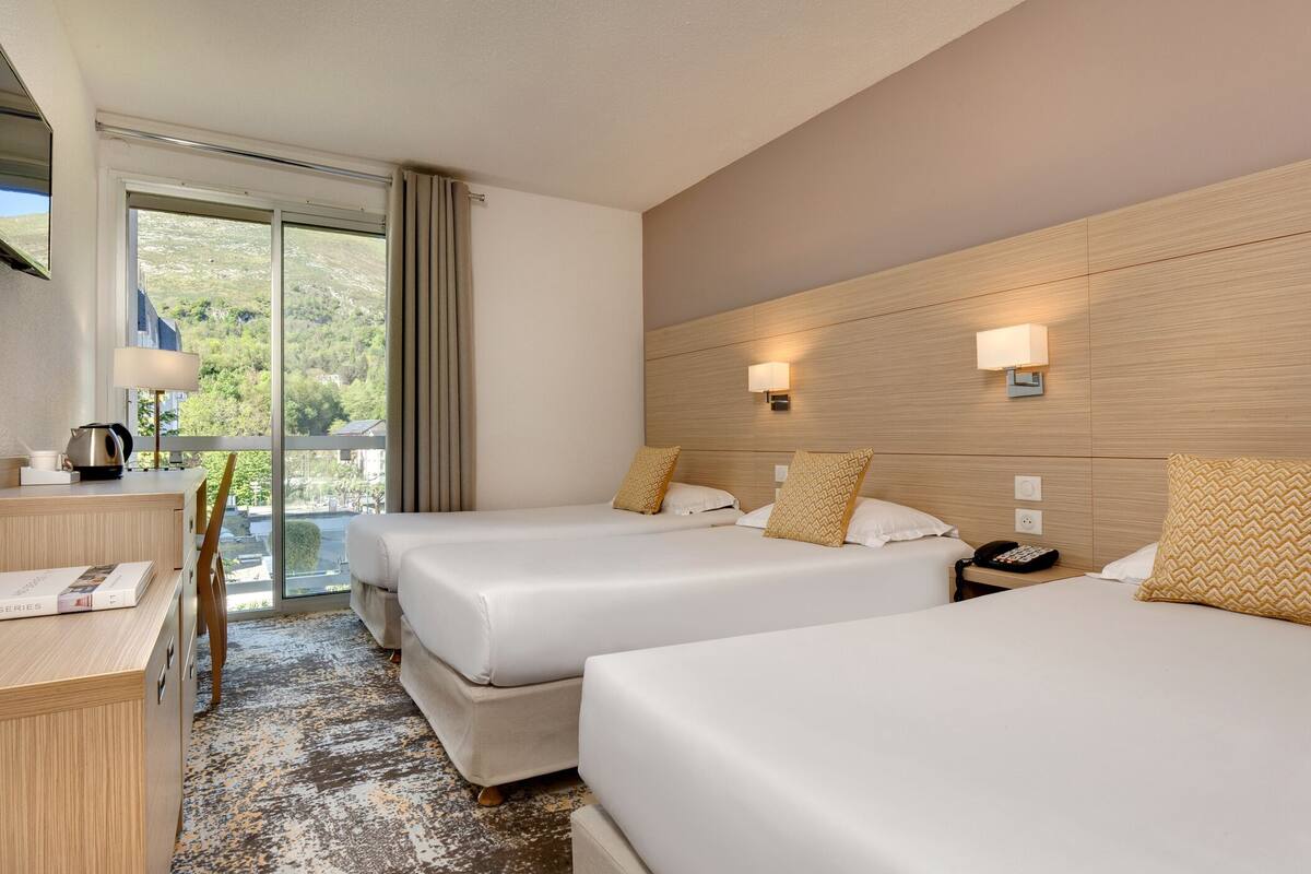 A hotel room with two beds, a desk, and a view of the outdoors.