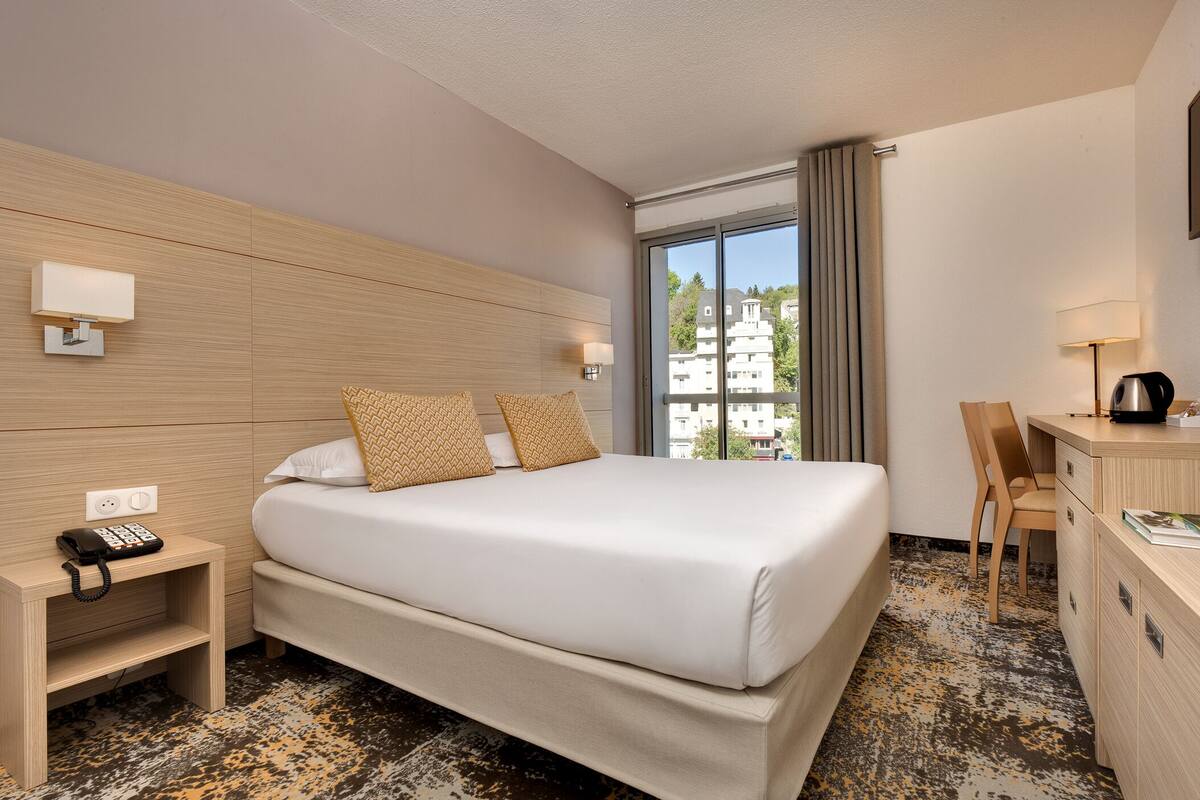 A hotel room with a large bed, bedside tables, a desk, and a view of buildings through the window.