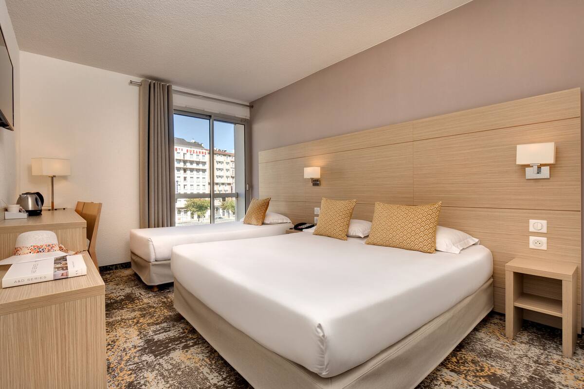 A hotel room with two beds, a desk, a chair, and a window with a view of buildings.