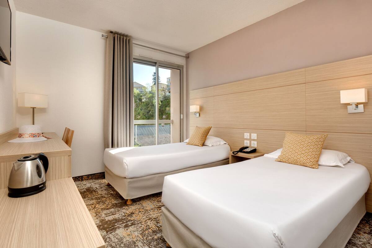 A hotel room with two beds, a desk with a kettle, a lamp, and a view of greenery outside.