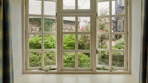 A bay window with multiple panes, offering a view of a garden with various plants and a brick building in the background. - The Arundell  (Lifton)