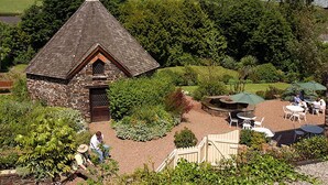 A stone cottage with a triangular roof, surrounded by lush greenery and a patio area with outdoor seating. - The Arundell  (Lifton)