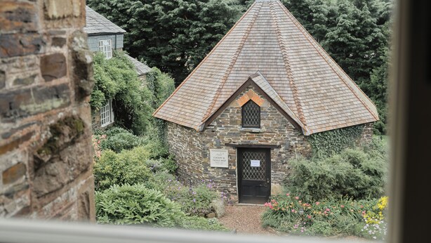 A stone building with a steeply pitched roof, surrounded by lush greenery and a sign indicating it is a craft shop. - The Arundell  (Lifton)