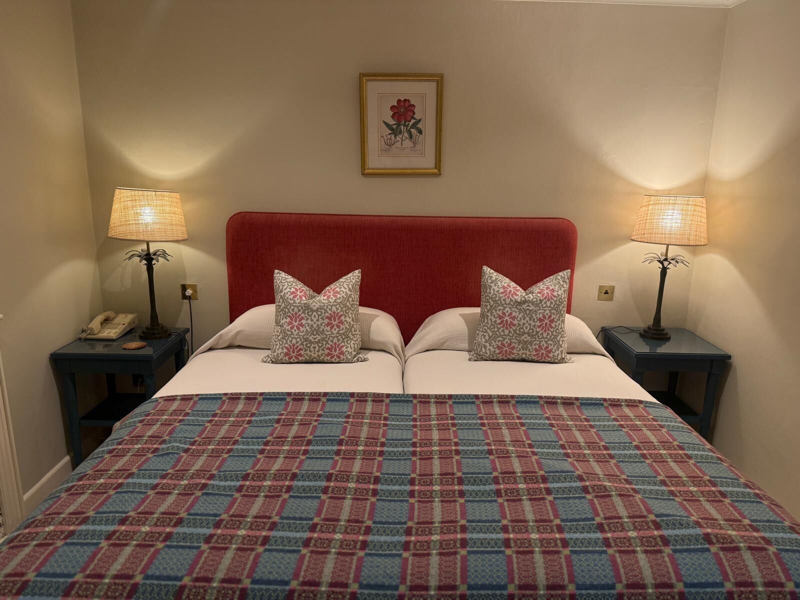 Una cama doble con cabecera roja, dos almohadas estampadas, una obra de arte floral en la pared, dos mesitas de noche con lámparas y una colcha estampada.