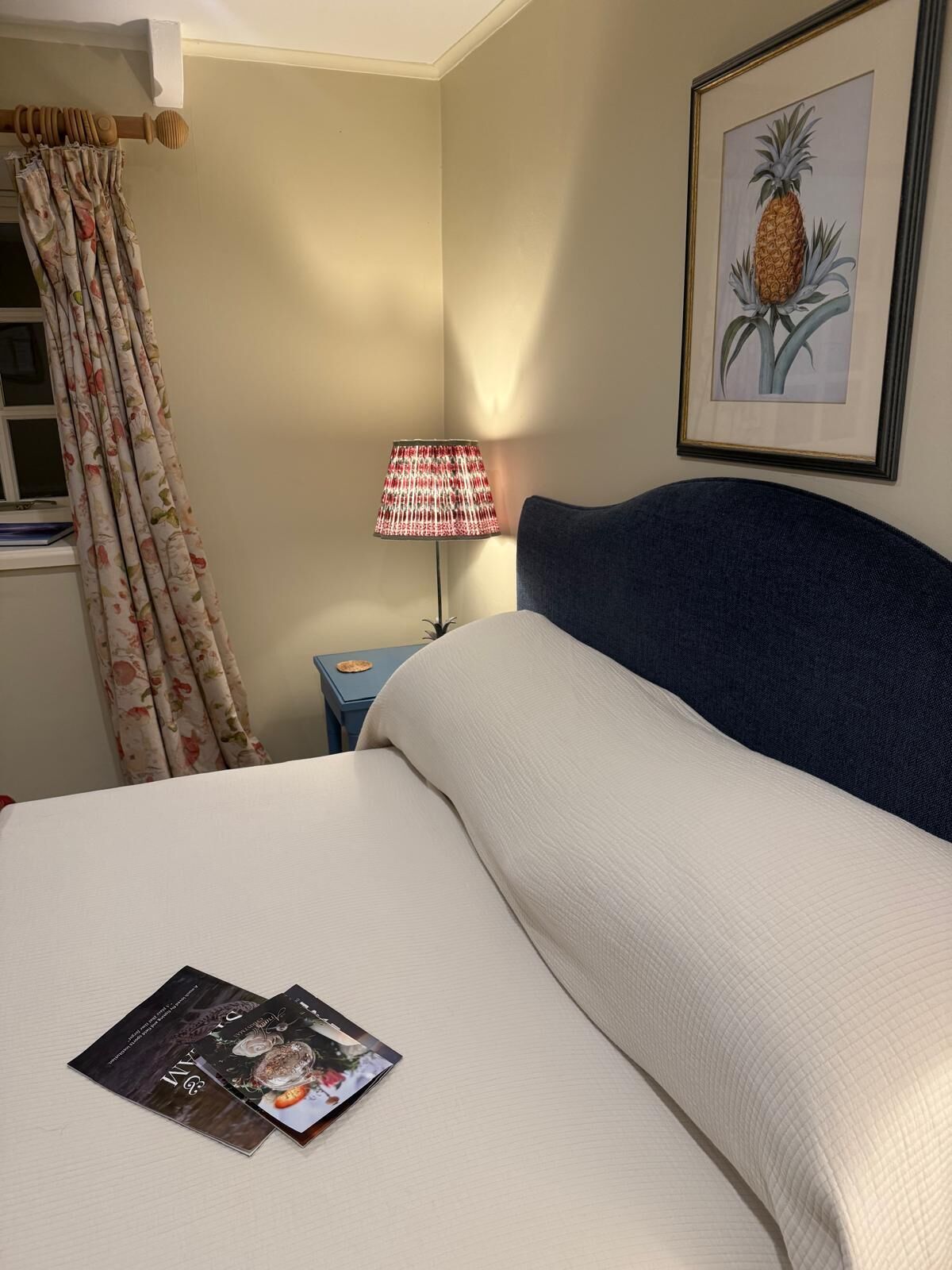 Ein ordentlich bezogenes Bett mit Kissen und einer Zeitschrift darauf. Auf einem kleinen Nachttisch neben dem Bett steht eine Lampe mit einem rot-weißen Lampenschirm. An der Wand hängt ein gerahmtes Bild von einer Ananas.