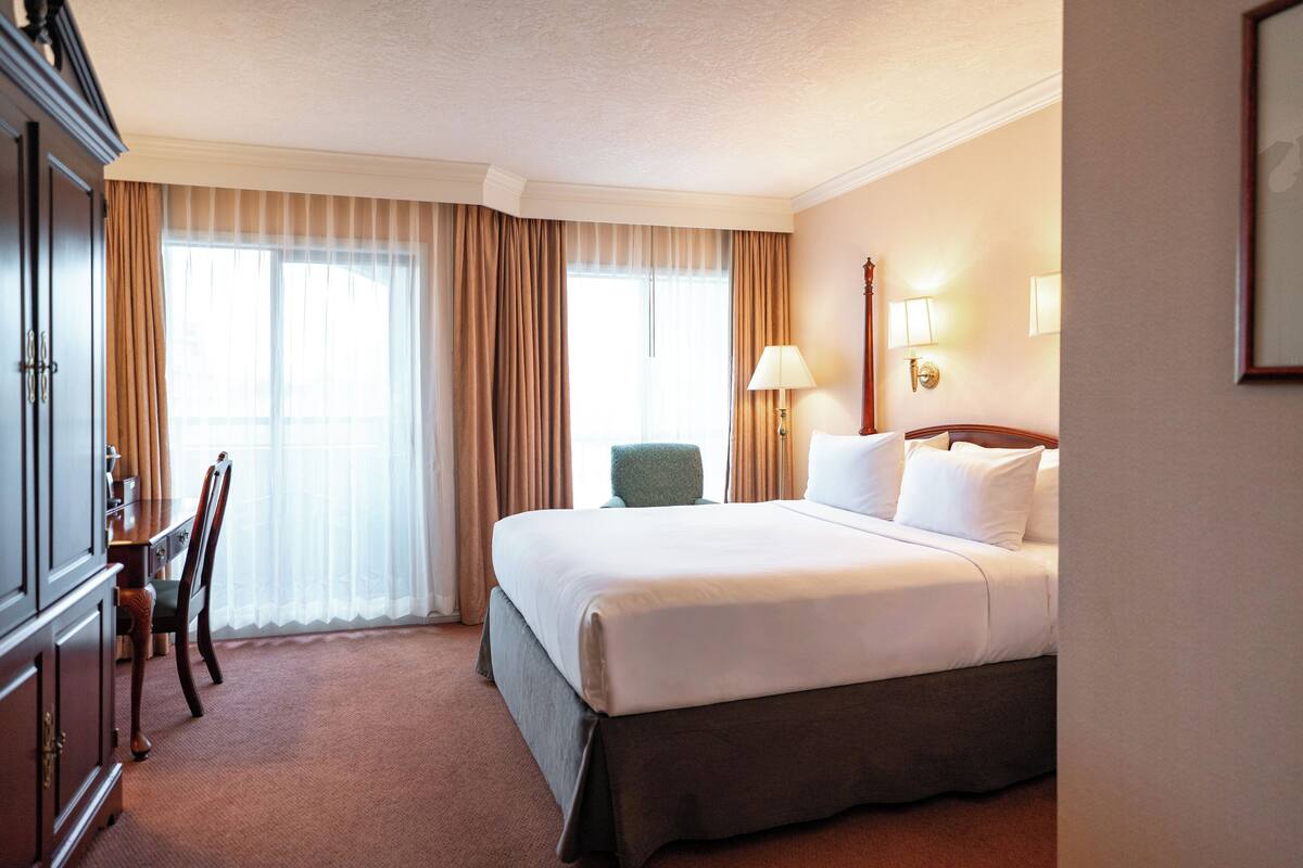 A hotel room with a large bed, a desk with a chair, a lamp, and a window with curtains.