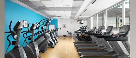 A gym with treadmills and elliptical machines, wooden flooring, and large windows.