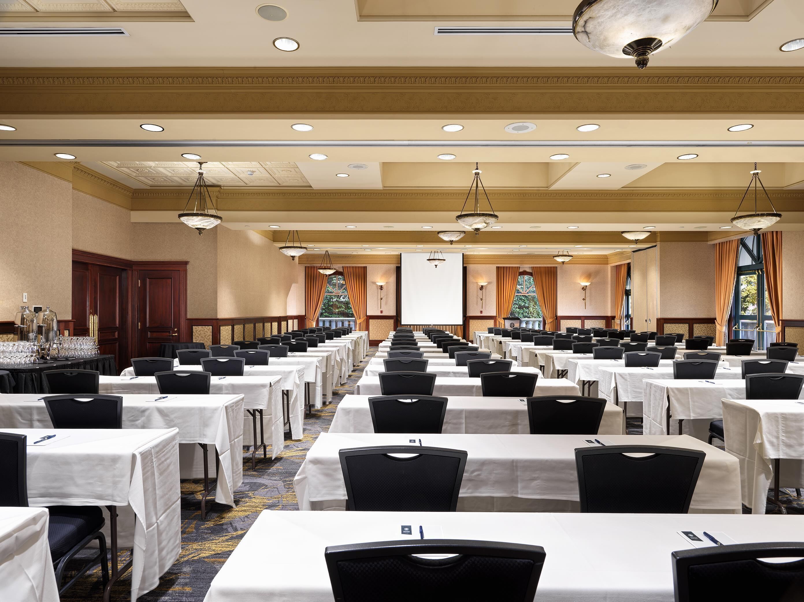 A large conference room with rows of tables and chairs, a projector screen, and a large window with curtains.