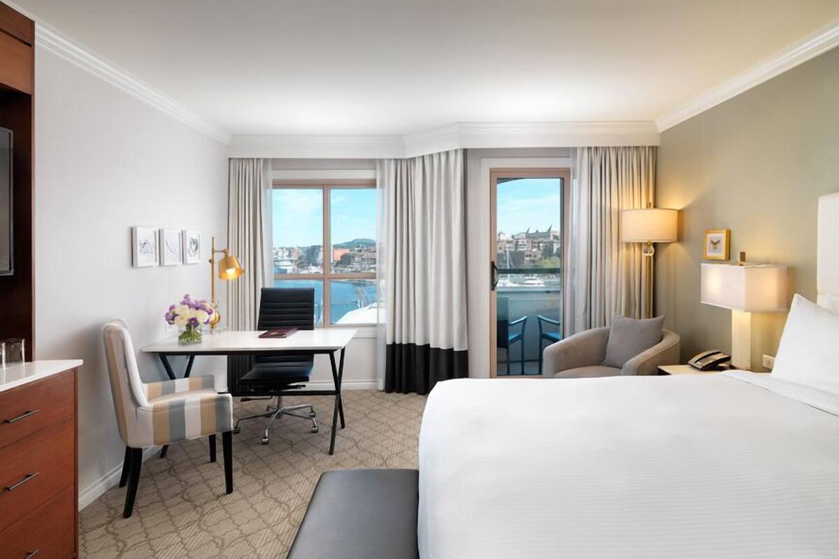 A hotel room with a bed, a desk with a chair, a small table with a vase of flowers, a sofa, and a view of the city through large windows.