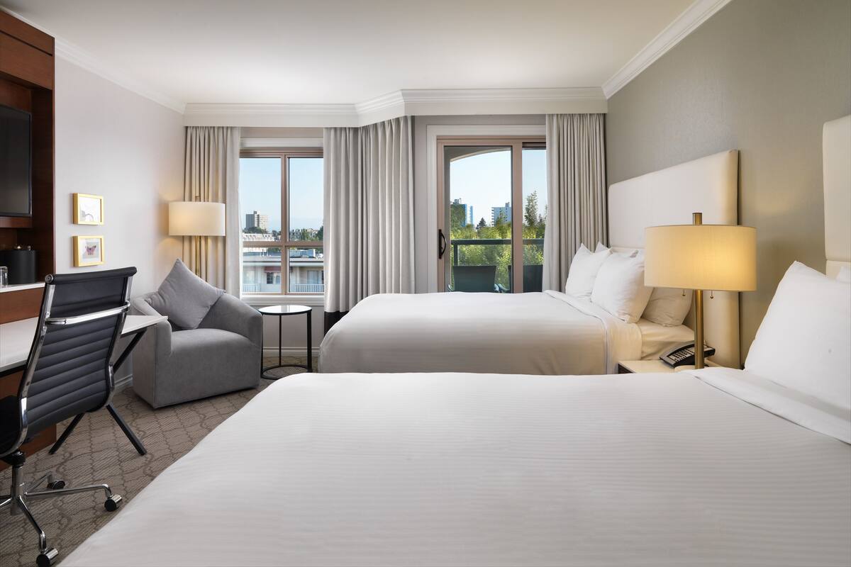 A hotel room with two beds, a desk, a chair, a lamp, and a view of the city through large windows.