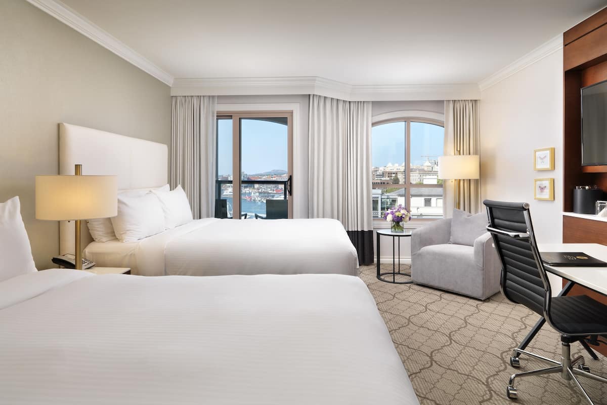 A hotel room with two beds, a desk, a chair, a TV, and a view of the city through large windows.