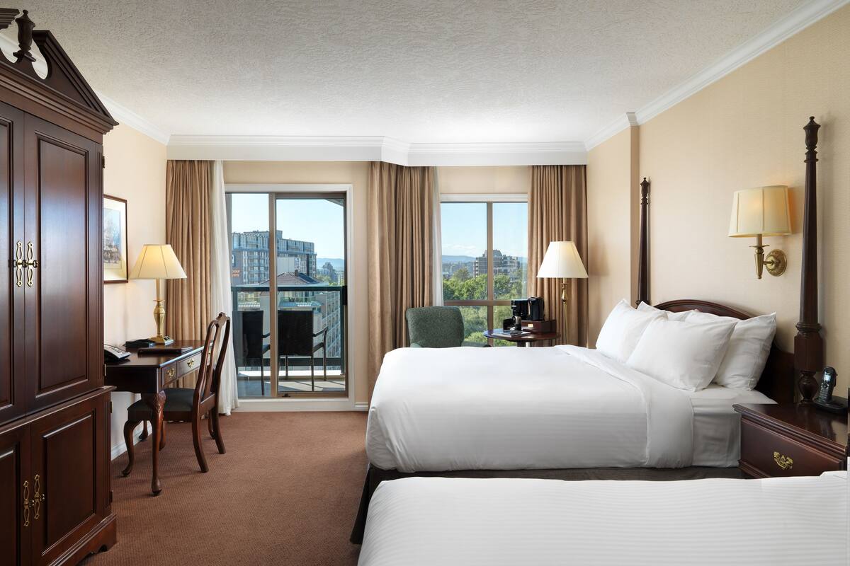 A hotel room with two beds, a desk, a chair, a lamp, and a view of the city.