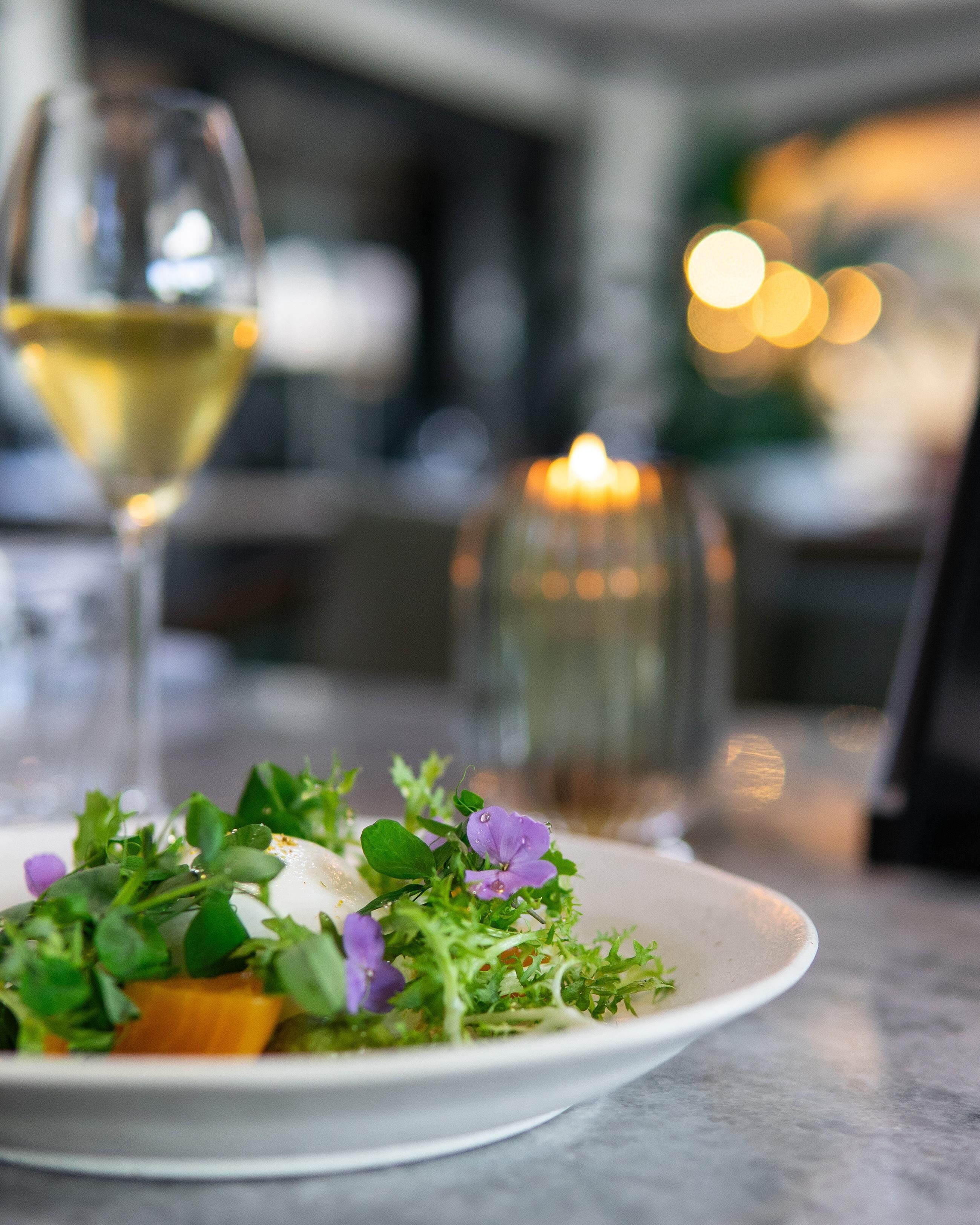 A plate of food with greens and purple flowers, a glass of white wine, and a candle in the background.