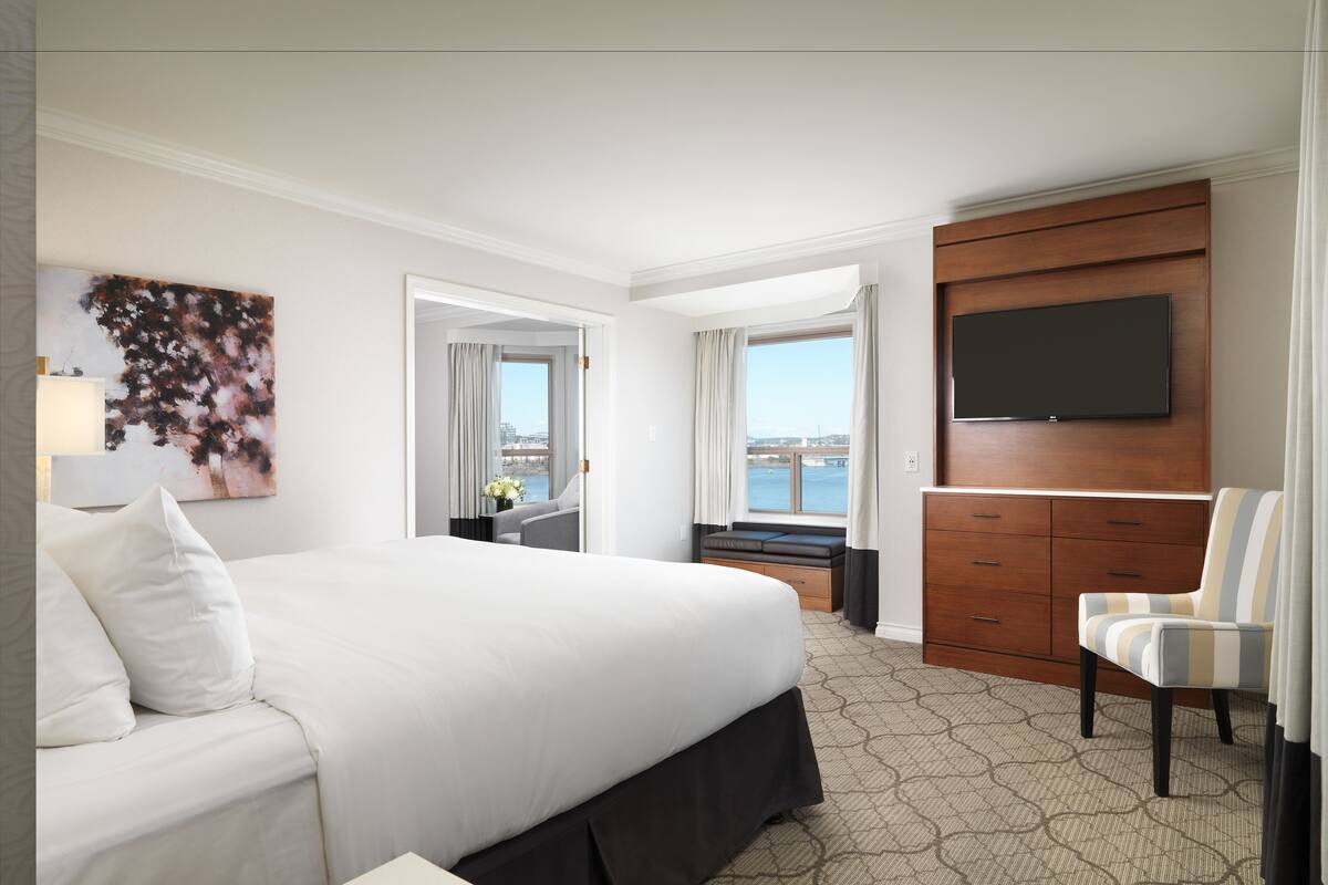 A hotel room with a large bed, a TV mounted on the wall, a seating area with a view of the water, and a window with curtains.
