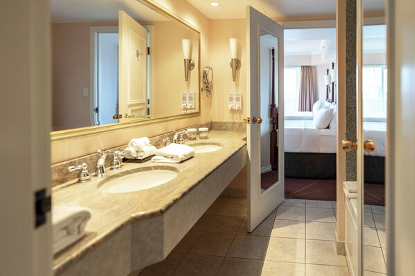 A hotel bathroom with two sinks, a large mirror, and a view into a bedroom with a bed and curtains.