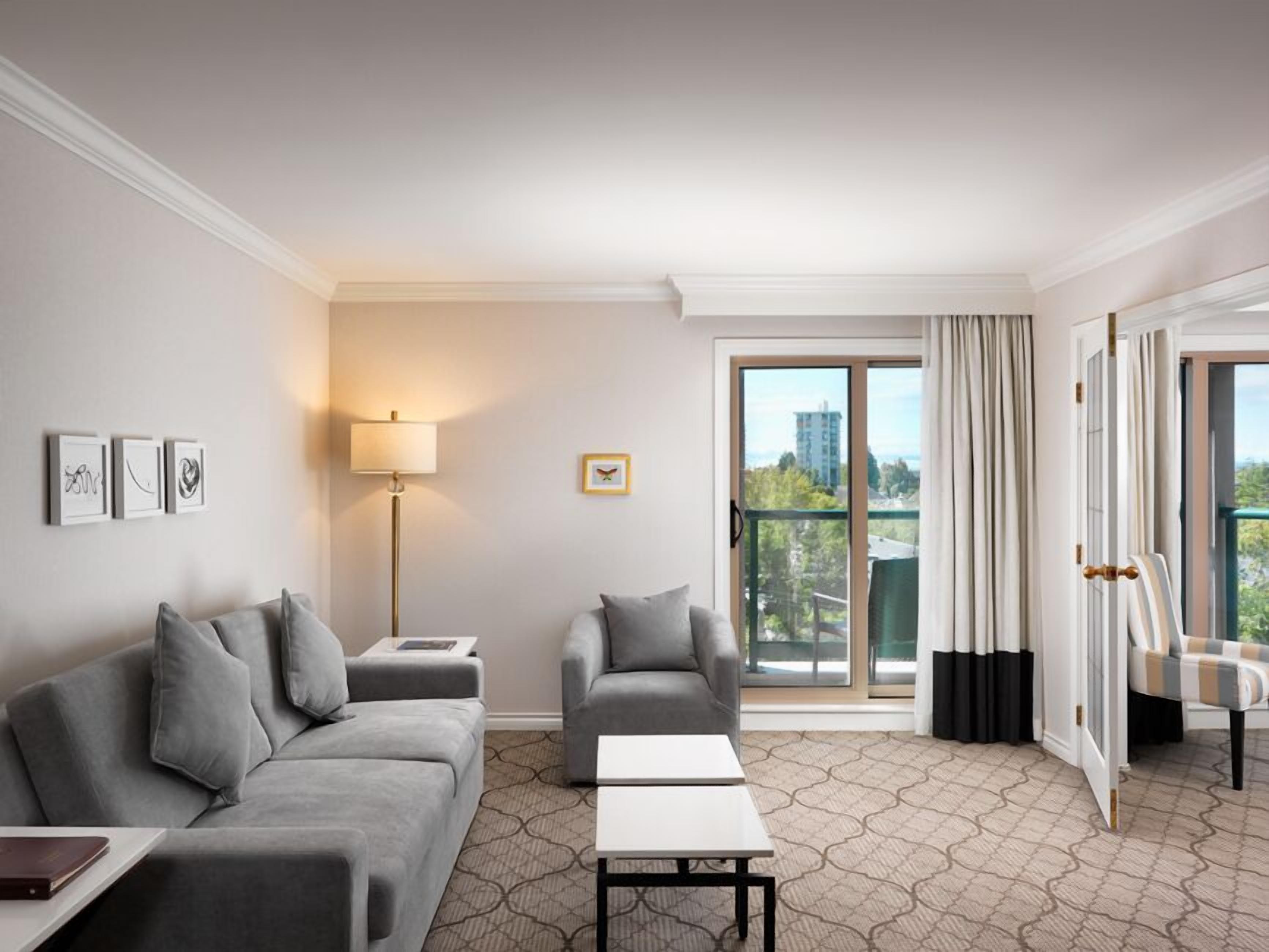 A modern hotel room with a grey sofa, a smaller armchair, a white coffee table, and a floor lamp. There is a large window with a view of buildings and trees.