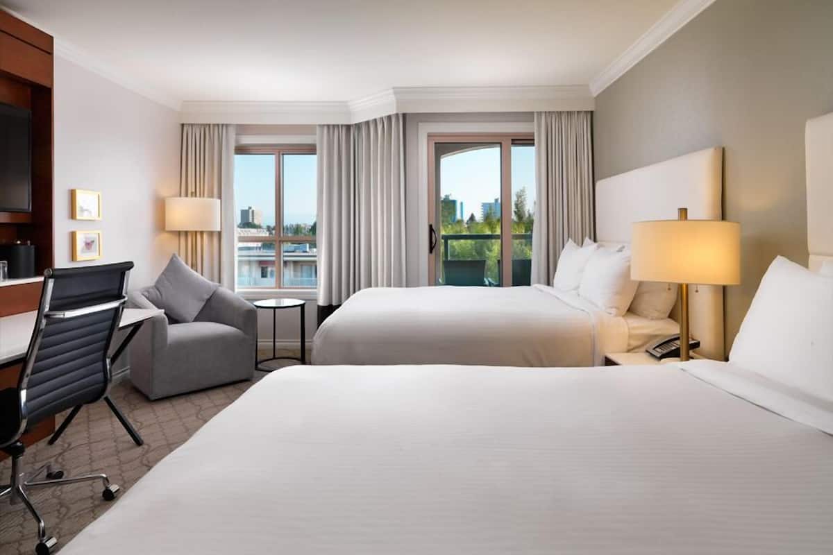 A hotel room with two beds, a desk, a chair, a lamp, and a view of the city.