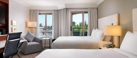 Habitación de hotel con dos camas, un escritorio, una silla, una lámpara y vistas a la ciudad.