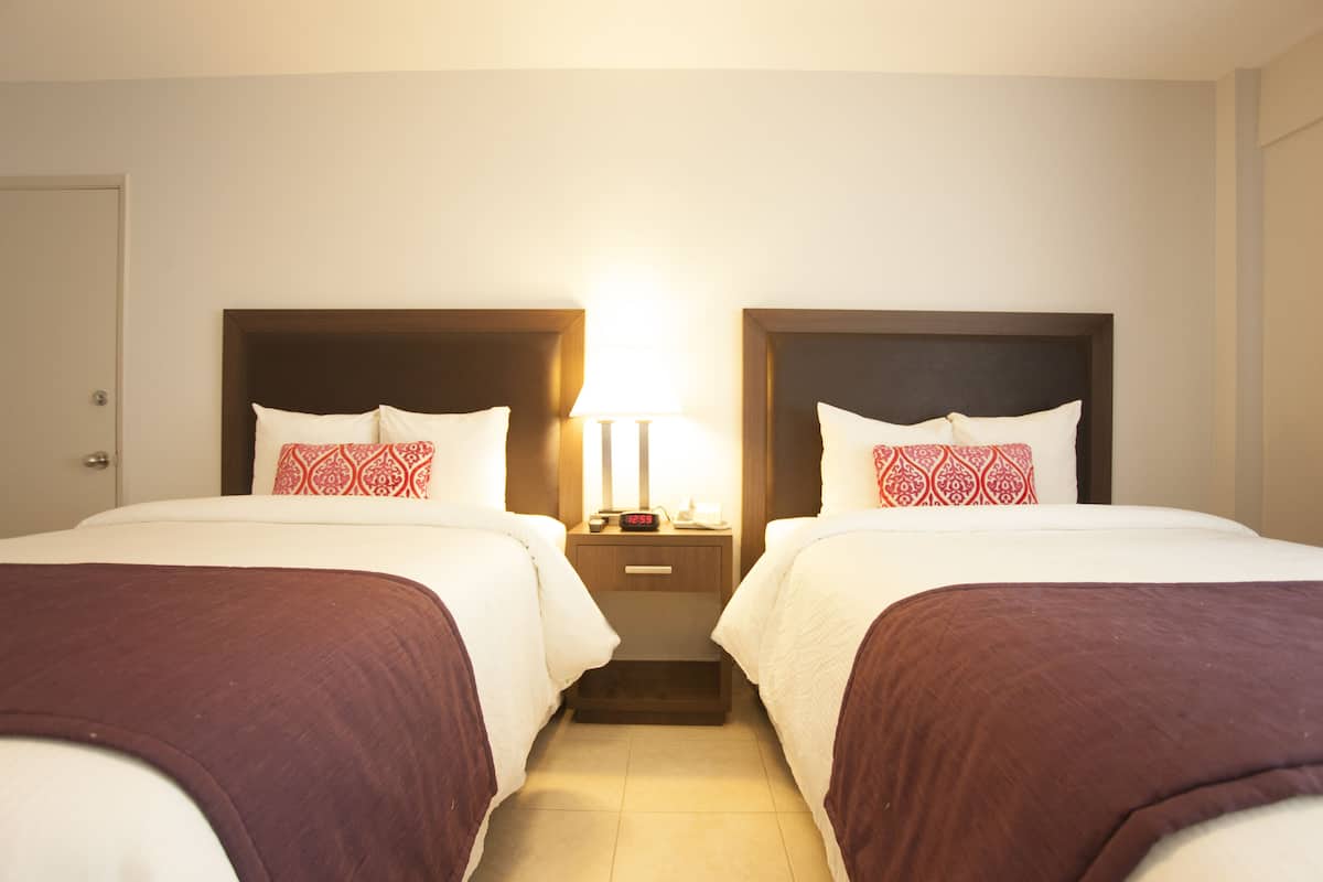 A hotel room with two beds, each with a wooden headboard, white bedding, and red patterned pillows. There is a nightstand between the beds with a lamp and a clock.