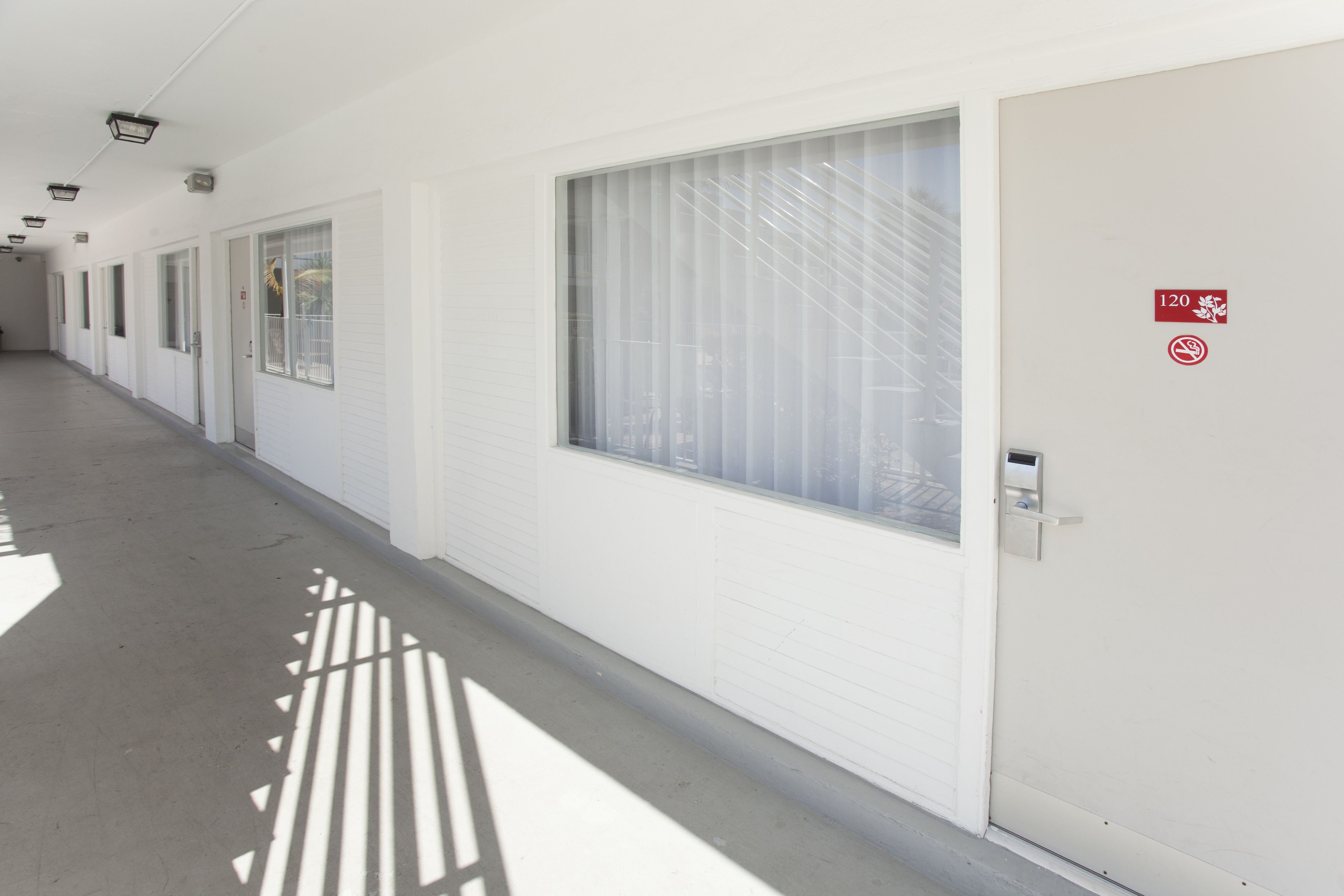 A long, narrow hallway with white walls and doors, each with a window. There is a no-smoking sign on one door.