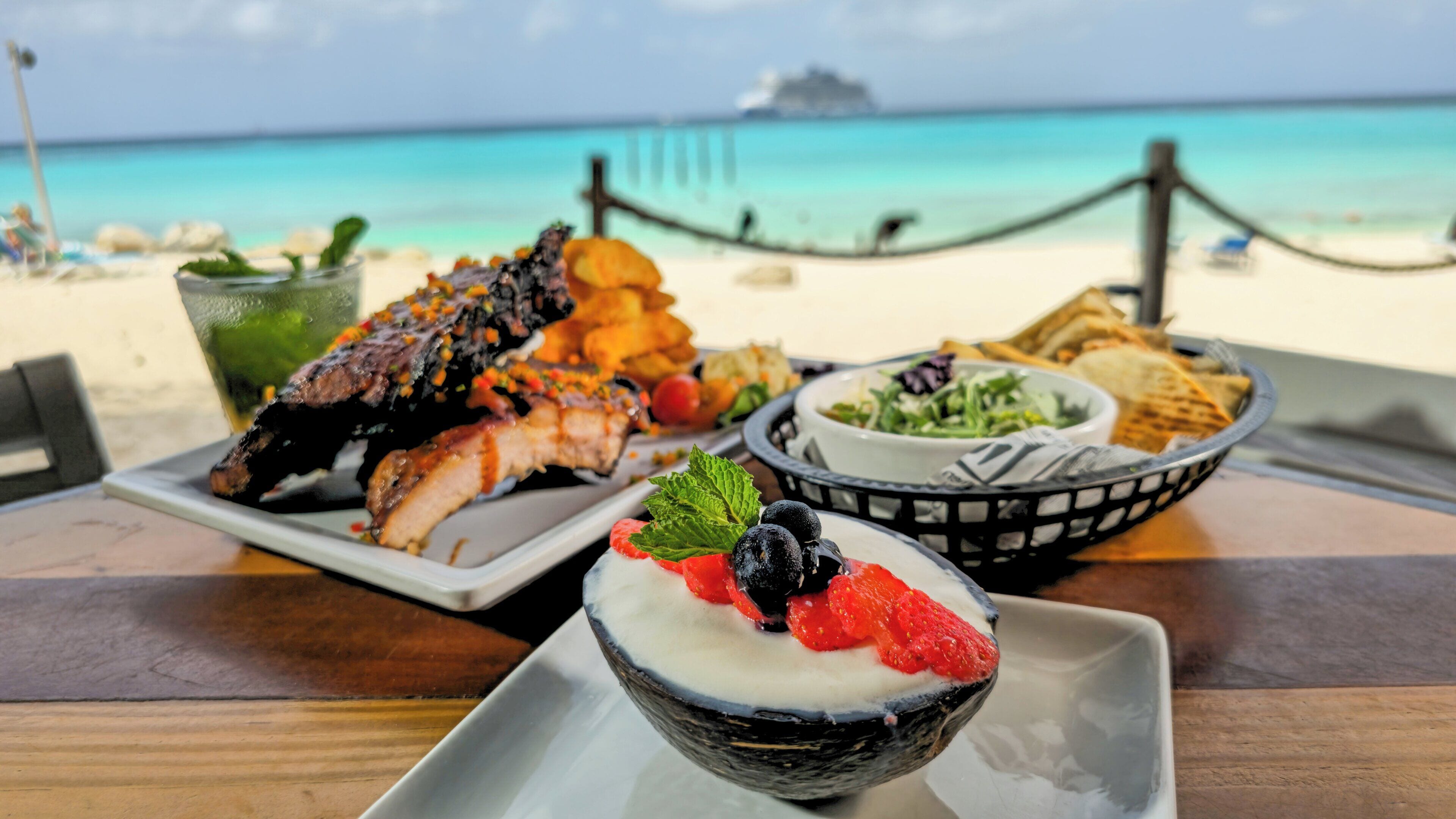 A dining table with a variety of dishes, including grilled meat, salad, and a dessert with berries, set against a beach backdrop.