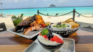 A dining table with a variety of dishes, including grilled meat, salad, and a dessert with berries, set against a beach backdrop.